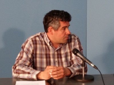 Un hombre con una camisa a cuadros está sentado en frente de un micrófono, probablemente en una conferencia o rueda de prensa. La pared al fondo es azul y hay una sombra en el suelo, lo que sugiere la presencia de luces.