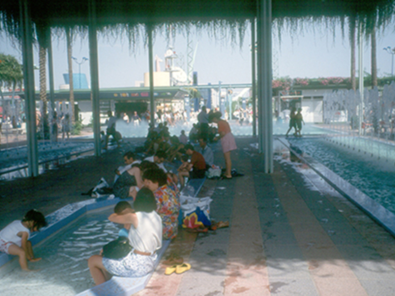Niños jugando en una piscina al aire libre con un techo de palmeras.