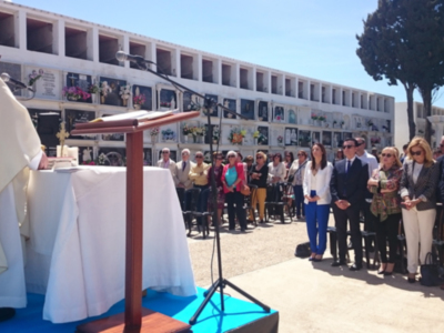 Una imagen de una ceremonia religiosa o formal con un sacerdote en el centro, frente a una mesa blanca y dorada. A su derecha, un grupo de personas vestidas formalmente observan la ceremonia. El fondo muestra edificios con ventanas y una línea de árboles en el horizonte. La imagen transmite un ambiente serio y ceremonial.