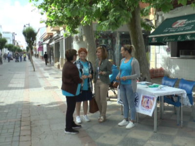 Tres mujeres conversan en un parque público, cerca de una mesa con una bandera y sombrillas azules. Detrás de ellas, hay tiendas y árboles con hojas verdes.