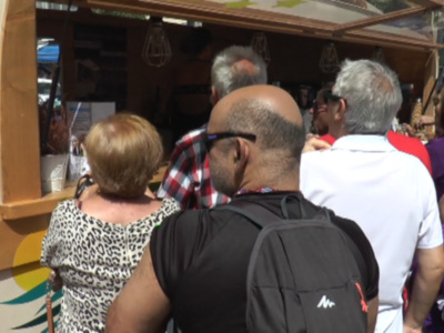 Un grupo de personas esperando en una fila frente a un food truck. La imagen muestra a varias personas con mochilas y ropa casual, incluyendo una mujer con un vestido de leopardo. El food truck tiene un diseño moderno con colores vivos y una iluminación que destaca su entrada. La escena parece ser en un evento al aire libre, posiblemente una feria o mercado.