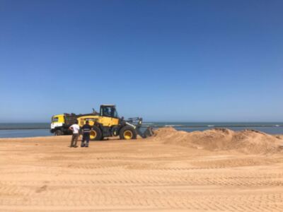 Una escena de construcción en la playa con un tractor amarillo y dos personas trabajando. El cielo es azul claro, el suelo es arenoso y hay grandes montículos de arena alrededor.
