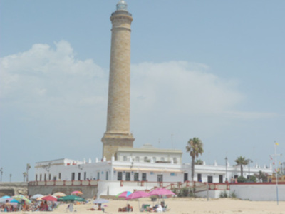 Una torre de mampostería en un entorno costero con playas y palmeras. La torre es alta, con una base ancha que se ensancha hacia arriba y termina en un ático. En la base de la torre, hay una estructura blanca con ventanas y una terraza. La playa está llena de sombrillas y personas disfrutando del sol. El cielo es claro con algunas nubes dispersas, indicando buen tiempo.