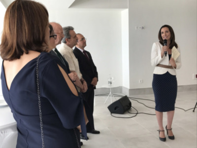 Una mujer con un vestido blanco y falda azul con puntos habla en una micrófono mientras tres hombres observan. El ambiente es formal, con un fondo blanco y equipo de sonido visible.