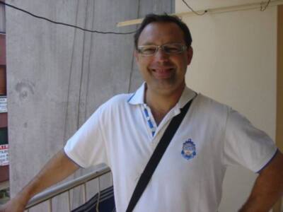 Un hombre con una camiseta blanca y un cinturón negro, posando frente a una pared de hormigón con una raya horizontal.