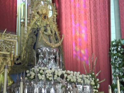 Una estatua religiosa dorada con un manto de plata, rodeada por flores blancas y velas encendidas. El fondo es un muro rojo con una decoración floral en la parte superior derecha.