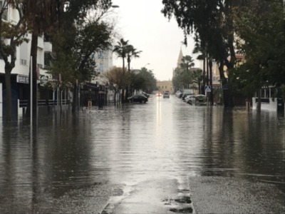 Calle inundada con vehículos y edificios al fondo, reflejo de lluvia en el agua.
