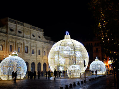 Escena nocturna de una plaza con luces navideñas en forma de esferas iluminadas. En el fondo, una estructura arquitectónica con ventanas y detalles decorativos. Personas caminando por el área iluminada, creando un ambiente festivo y navideño.