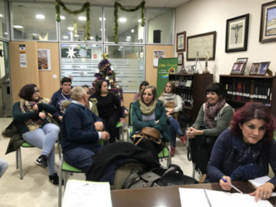 Una sala de espera con personas sentadas en sillas y una persona escribiendo en un libro. Hay decoraciones navideñas, como una araña de Navidad y un árbol en el fondo. La pared está decorada con cuadros y una estatua de un crucificado en la esquina.