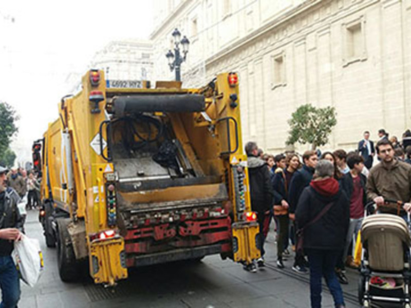 Una camioneta de limpieza urbana amarilla con una gran máquina de recogida de basura en la parte frontal, estacionada frente a un edificio blanco. La imagen muestra a varias personas caminando y esperando, algunas con carros de bebé. El entorno parece ser una calle urbana con árboles y edificios al fondo.