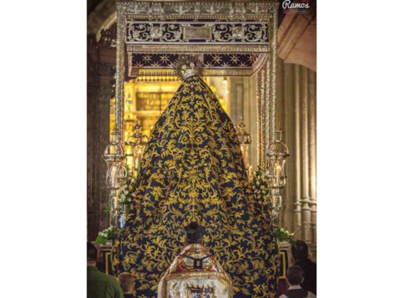 Una figura venerada en un altar de iglesia, envuelta en una manto dorado con detalles florales. La escena está iluminada por luces de cristal, creando un ambiente sagrado y reverente.