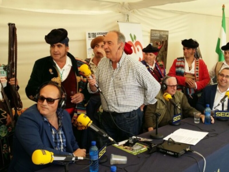 Se observa a un grupo de personas en una conferencia de prensa. Algunos están vestidos con trajes tradicionales y otros con ropa casual. En el centro hay un hombre mayor con barba, hablando mientras otros asisten a la conferencia. En el fondo se pueden ver banderas y un cartel informativo. La mesa está equipada con micrófonos, auriculares y otros dispositivos de transmisión.