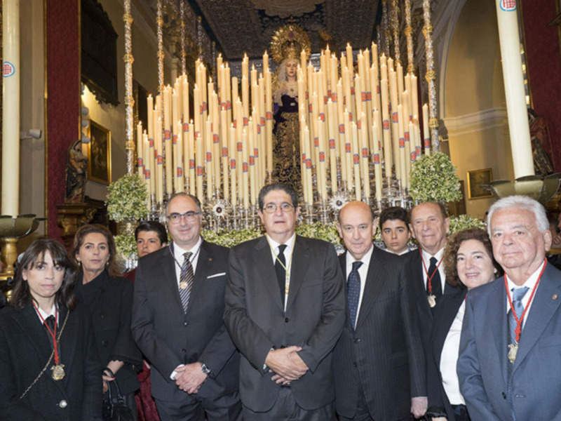 Una imagen de una ceremonia religiosa en un templo, con una estatua central y velas encendidas. La gente está vestida formalmente, con chaquetas y corbatas, posando para la foto. La decoración incluye detalles dorados y una gran imagen en el fondo.