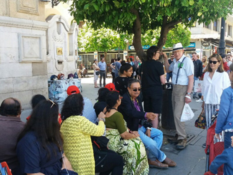 Grupo de personas sentados y caminando en una plaza urbana con árboles y edificios al fondo.