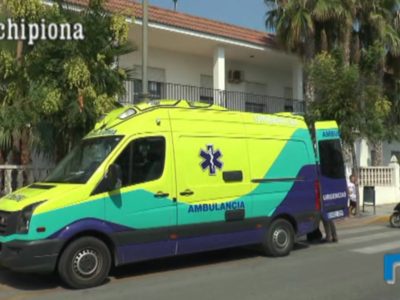 Un ambulancia verde y azul con el logo de la Cruz Roja, estacionada en una calle con palmeras y edificios blancos.