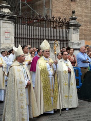 Un grupo de religiosos vestidos con hábitos y capas blancas, posiblemente sacerdotes, caminan por una calle con un edificio de ladrillo en el fondo. Algunos llevan insignias y medallas, indicando posiblemente su jerarquía o roles en la iglesia. La escena parece ser una procesión o ceremonia religiosa, con un ambiente formal y solemn.