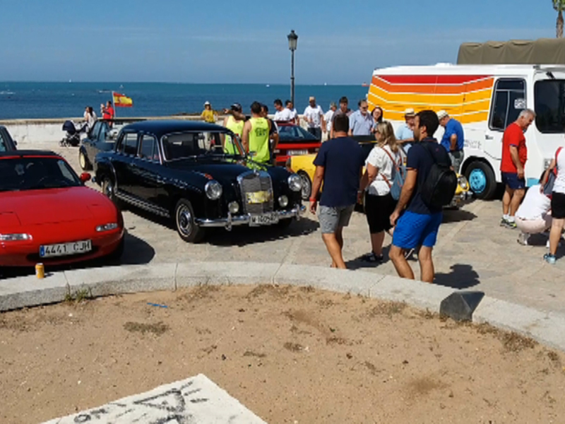 Se observa una escena en la playa con varios coches estacionados cerca del mar. Los vehículos son de diferentes modelos y colores, incluyendo un coche rojo, uno negro y otro con una combinación de colores. Al fondo se puede ver el horizonte del mar y algunas personas caminando por la orilla. La atmósfera es relajada, típica de un día soleado en la playa.