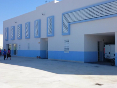 Fachada de un edificio industrial con ventanas azules y puertas abiertas. Un vehículo blanco está estacionado en la entrada. Personas caminan alrededor del edificio.