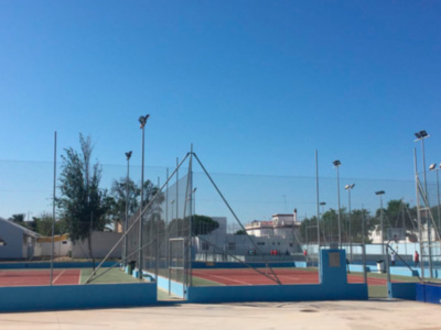 Cancha de tenis con redes y postes altos, bajo un cielo azul claro. La cancha está rodeada por una cerca y hay árboles en el fondo.