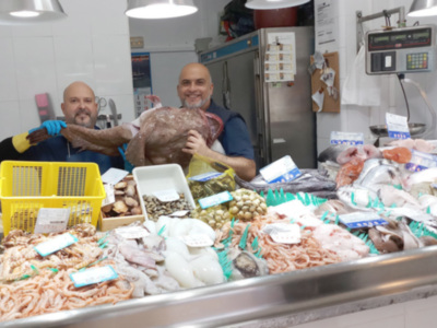 Dos hombres sostienen un pollo en una tienda de pescados y mariscos. En el fondo, se pueden ver refrigeradores y un mostrador con diversos productos frescos. La imagen destaca la variedad de mariscos y pescados disponibles en el establecimiento.