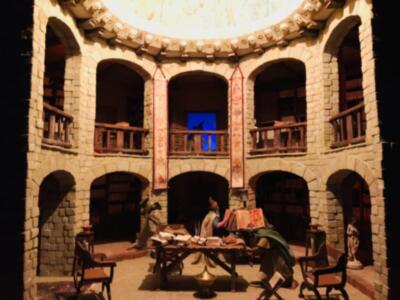 Un interior medieval de piedra con arcos y balcones, donde dos personajes se sientan en una mesa baja. La atmósfera es histórica y dramática, con detalles como un techo dorado y una figura en la parte superior.