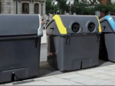 Tres contenedores de reciclaje en una calle con edificios y árboles al fondo.