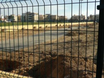 Un terreno despejado con una estructura de hormigón en el fondo, probablemente un estadio o campo deportivo. Se puede ver una cerca de metal en primer plano, lo que sugiere que el terreno está protegido o limitado. En el fondo, se distinguen edificios urbanos bajo un cielo despejado.
