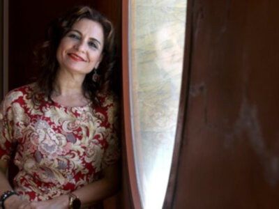 Una mujer con cabello largo y ondulado, vistiendo un vestido rojo con patrones florales, sonríe mientras se encuentra en una habitación con un mapa de fondo.