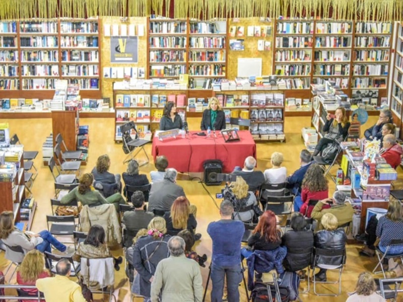 Una biblioteca con estanterías llenas de libros. Un grupo de personas sentadas en sillas alrededor de una mesa con libros y un hombre tocando la viola. La pared tiene carteles informativos y una pizarra. El suelo es de madera y hay cortinas en el fondo.