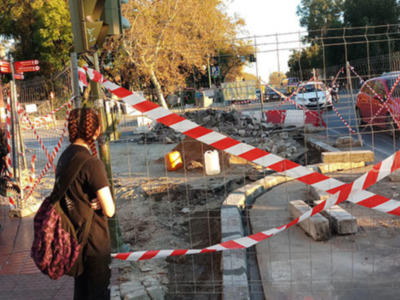 Un hombre y una mujer observan con atención un área de construcción bloqueada por barreras rojas y blancas, mientras una camioneta de trabajo circula en el fondo.