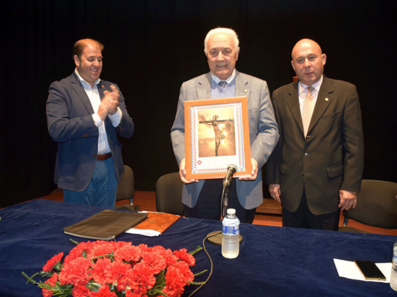 Tres hombres en trajes celebran una ceremonia, uno sostiene un marco con una imagen religiosa.