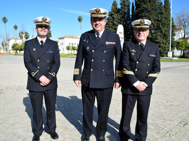 Tres oficiales de la marina en uniforme militar, posando frente a un edificio blanco con árboles verdes y cielo azul.