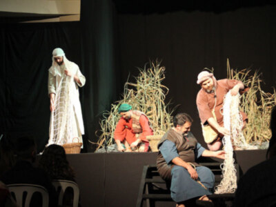 Actores en escena representando una escena de pesca, con un personaje central en túnica blanca y otros manipulando redes de pesca.