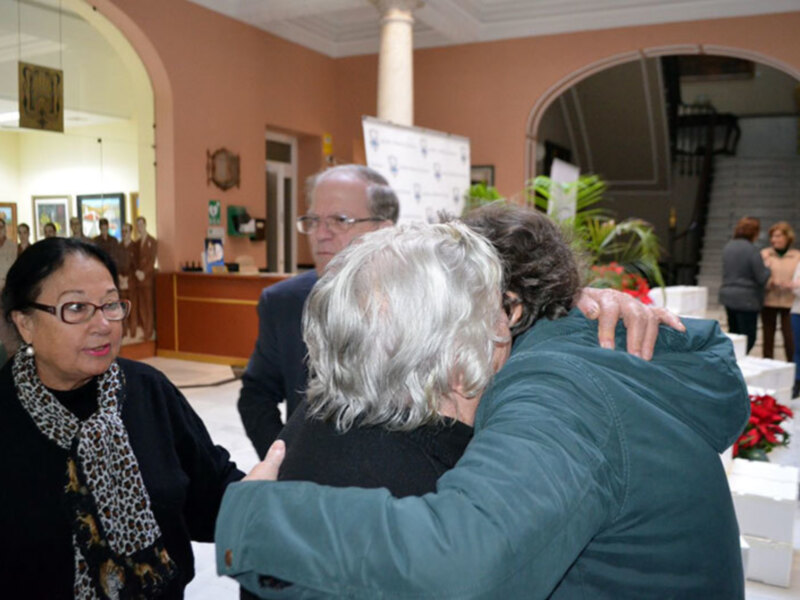 Un grupo de personas se abraza en un salón con decoración elegante, incluyendo flores y una columna central. La atmósfera es amistosa y festiva, probablemente en un evento organizado.