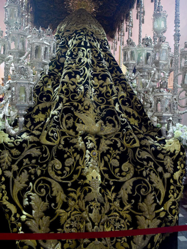 Imagen de un manto bordado con motivos florales y geométricos en tonos dorados sobre fondo negro. El manto parece ser parte de una vestimenta religiosa o ceremonial, posiblemente de un traje de sacerdote. El fondo muestra una estructura decorativa, posiblemente parte de un altar o edificio religioso.