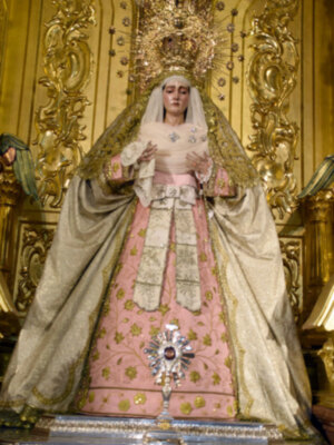 Imagen de la Virgen María vestida con un manto rosa y dorado, con una corona de diamantes y un escudo en la frente, en una iglesia con decoración dorada en el fondo.