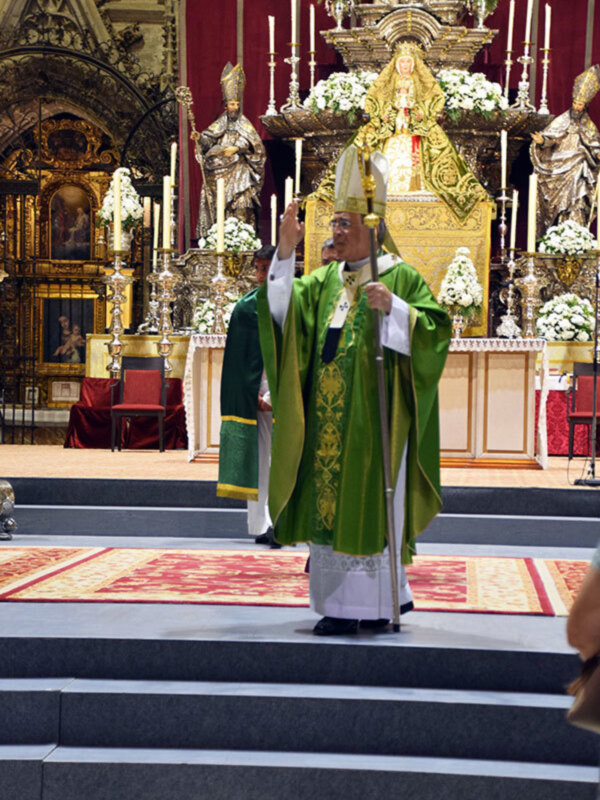 Imagen de una iglesia con un altar en el centro, donde un sacerdote viste vestimentas religiosas y realiza una oración. Alrededor del altar hay flores blancas, velas encendidas y estatuas de figuras religiosas. La decoración es elaborada con detalles dorados y madera.