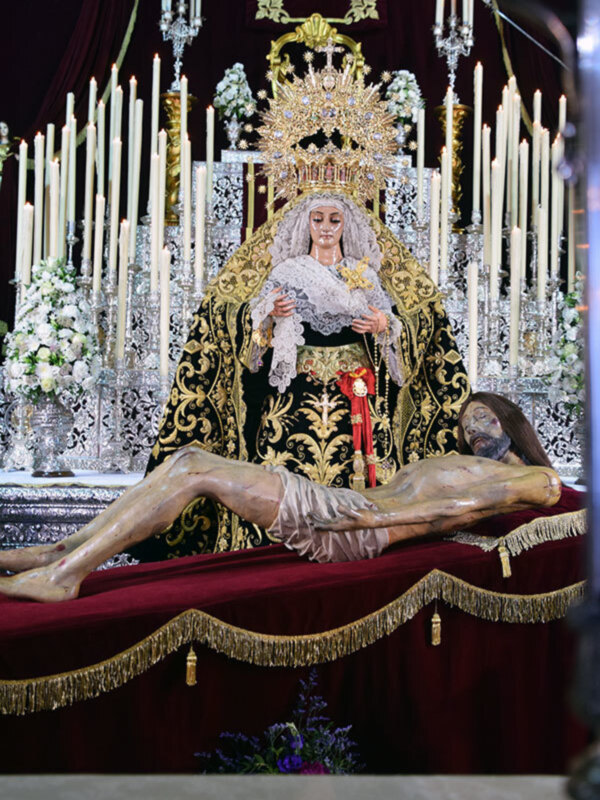 Una imagen de una Virgen María vestida con un manto blanco y coronada, sentada en un trono de oro. A su lado, una figura humana en posición de oración. Ambos están rodeados por velas altas y flores blancas en un entorno de lujo.