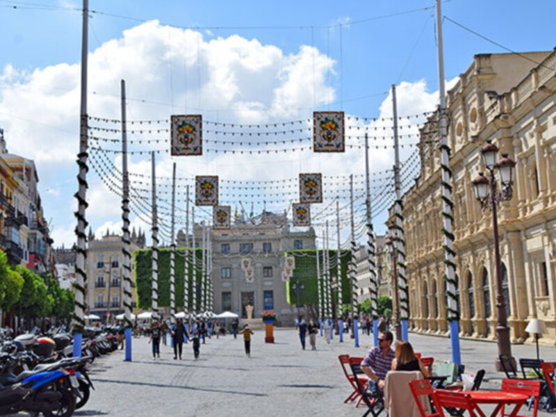 Plaza con edificios históricos, banderas colgadas en la red eléctrica, gente caminando y sillas rojas alrededor.