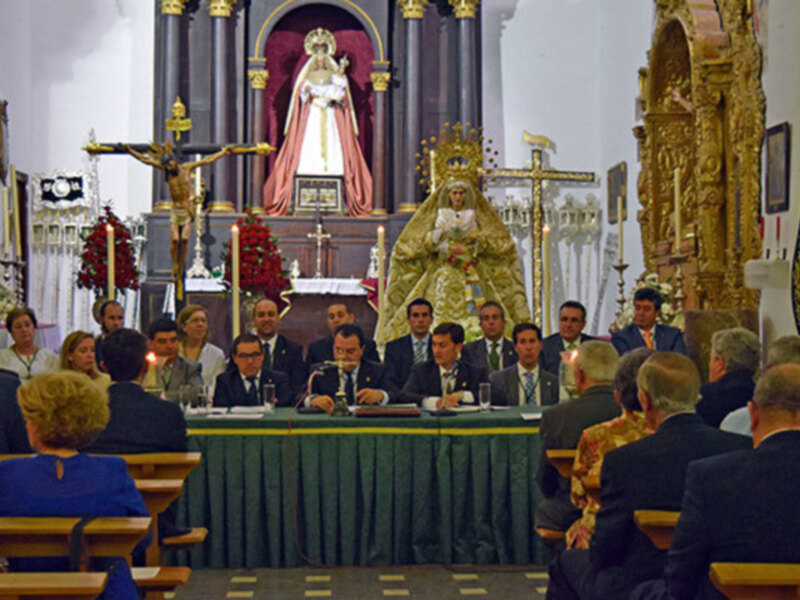 En una iglesia, un grupo de personas se reúne en un altar con una imagen dorada. La imagen central es rodeada por elementos decorativos y luces doradas. En la parte frontal, hay una mesa con sillas para los asistentes. La decoración incluye flores rojas y un crucifijo en la parte superior izquierda.
