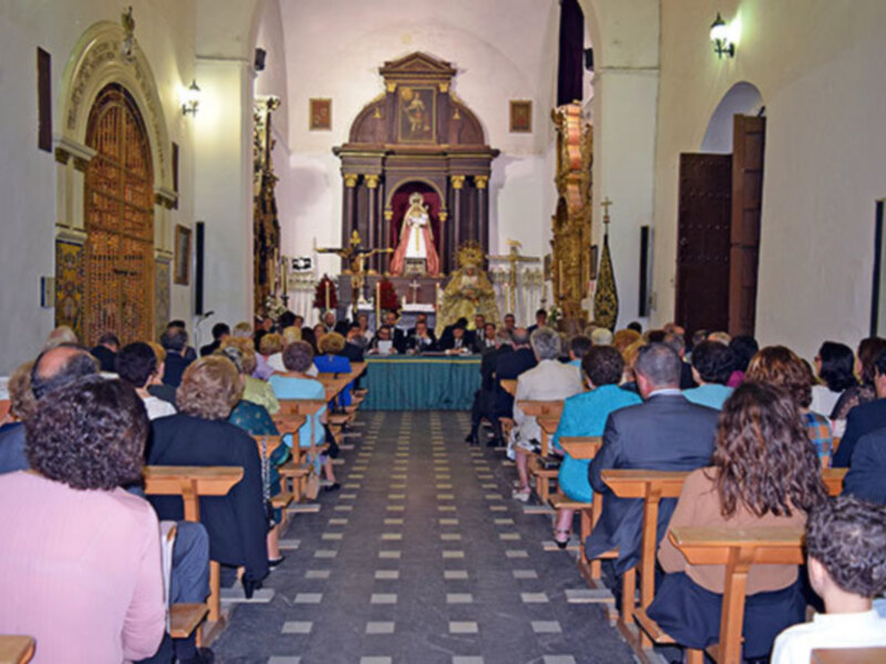 Interior de una iglesia con una imagen de la Virgen en el altar, asistentes sentados en bancas y una decoración arquitectónica en el fondo.