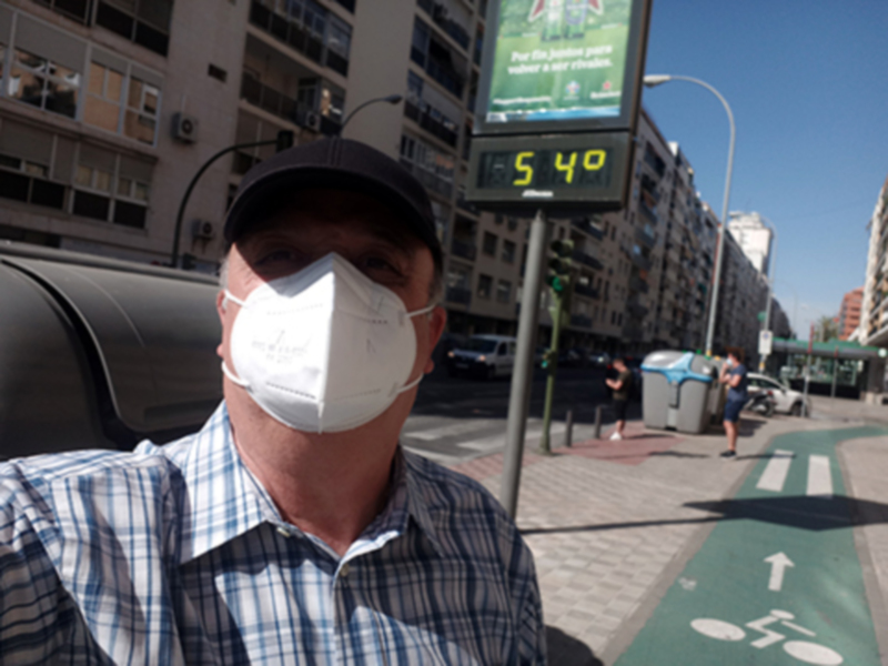 Un hombre con una mascarilla blanca y un gorra de béisbol está en la calle. A su lado, hay una señal de tráfico que muestra 5:40 y un camino peatonal con líneas blancas. En el fondo, se pueden ver edificios y una calle con coches pasando.