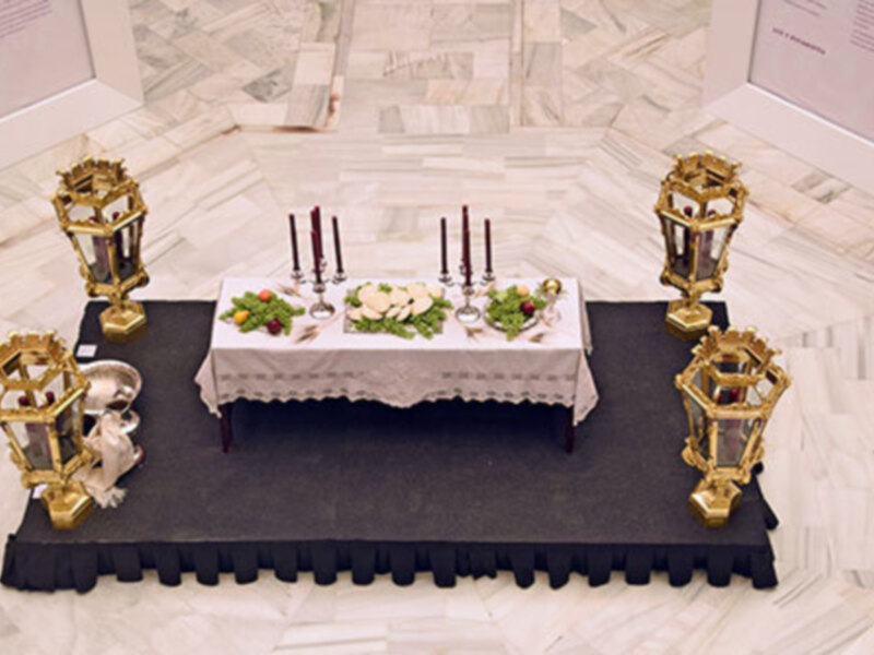 Un pequeño altar con velas encendidas, flores y un plato de pan en una mesa blanca. Alrededor, dos faroles dorados con figuras de ángeles y una alfombra negra. El fondo es un suelo de azulejos de color claro.