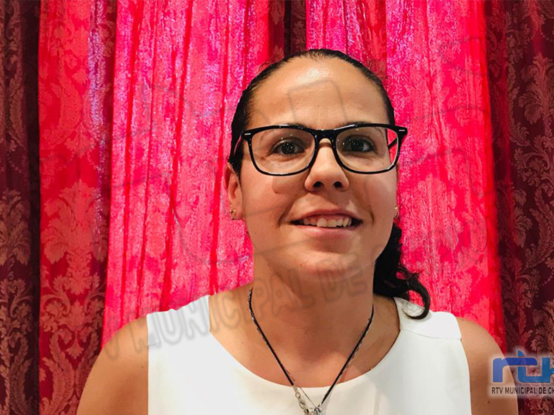 A una persona con cabello recogido en dos coletas, llevando gafas y un vestido blanco. El fondo es una cortina roja con detalles dorados, probablemente en un evento formal o ceremonial.