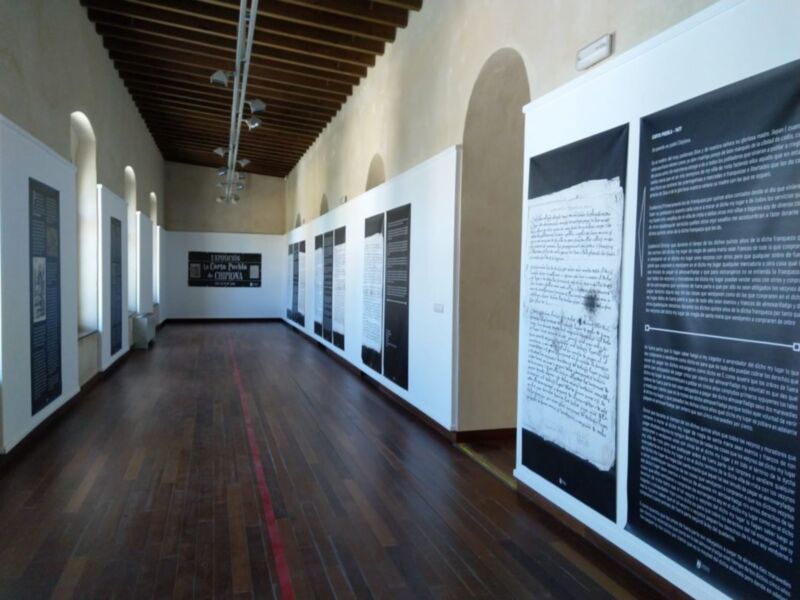 Salón de exposiciones con muros blancos y paneles informativos. Piso de madera, techo con vigas y luces de techo. Arcos en las paredes laterales.
