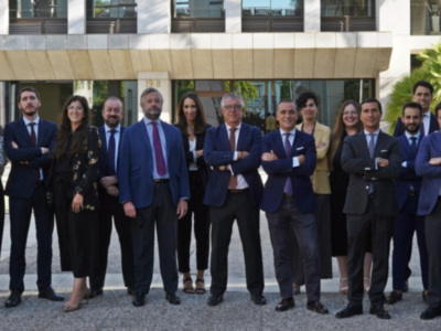 Una imagen de una fila de personas vestidas con trajes y chaquetas, posando frente a un edificio moderno. La escena está iluminada por el sol, con palmeras y árboles al fondo.