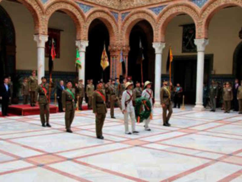 Una imagen de una ceremonia militar en un palacio con columnas y arcos. Los soldados están vestidos con uniformes militares, algunos portando banderas y otros con insignias. La escena es formal y ceremonial.
