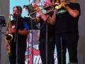Un grupo de músicos en un escenario, tocando saxofón y trompeta frente a una pantalla grande con imágenes de Coca-Cola.