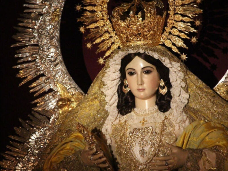 Imponente imagen de la Virgen con delicadas joyas y corona de oro, rodeada de luz y adoración.