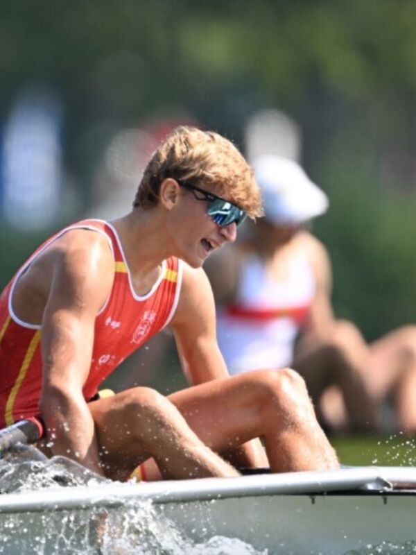 Un atleta de remo en acción, con una camiseta roja y amarilla, manejando un remo en el agua. La escena está al aire libre con otros atletas y un cielo despejado en el fondo.